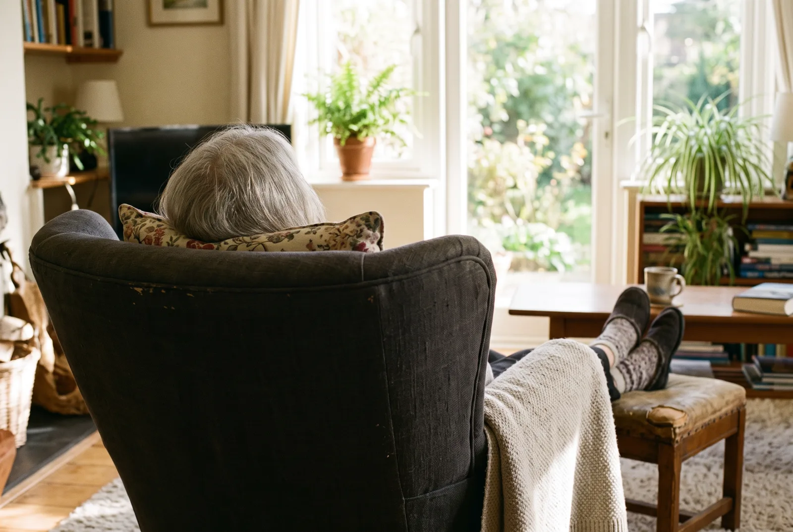Seen from behind a high-backed armchair, an older person rests by a sunny window with their feet up on an ottoman