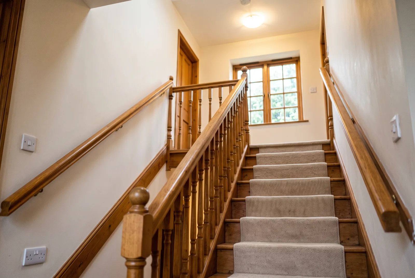 A bright, carpeted staircase in a family home with wooden handrails on both sides and a light switch at the bottom of the stairs
