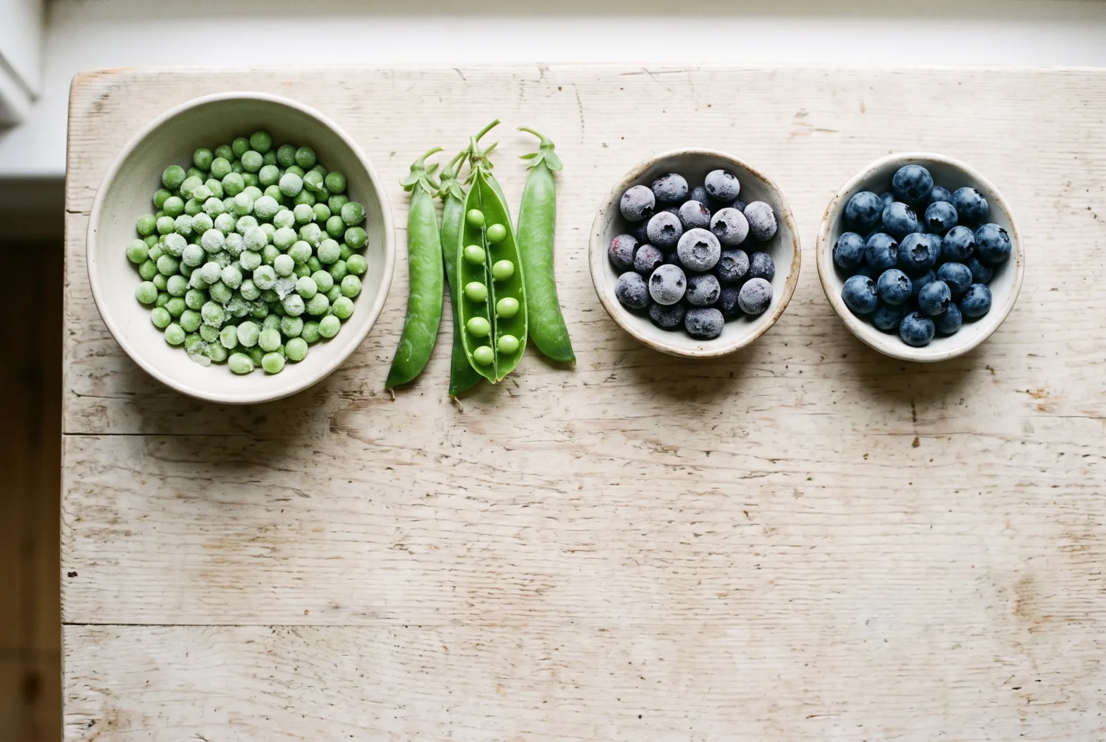 Bowls of frozen peas and frozen blueberries beside fresh pea pods and fresh blueberries on a pale wooden table