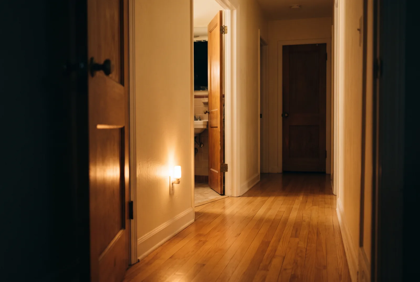 A quiet hallway at night softly lit by a small plug-in night-light, with a clear wooden floor leading to an open bathroom door