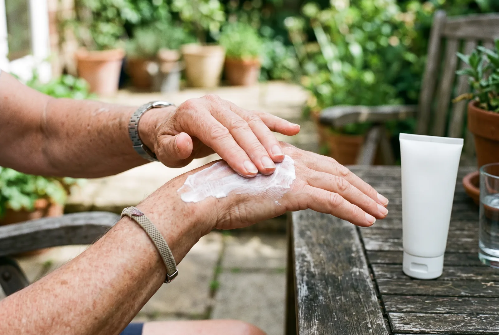 Older hands spreading white sunscreen lotion over the back of one hand on a garden patio, a plain white tube on the table nearby